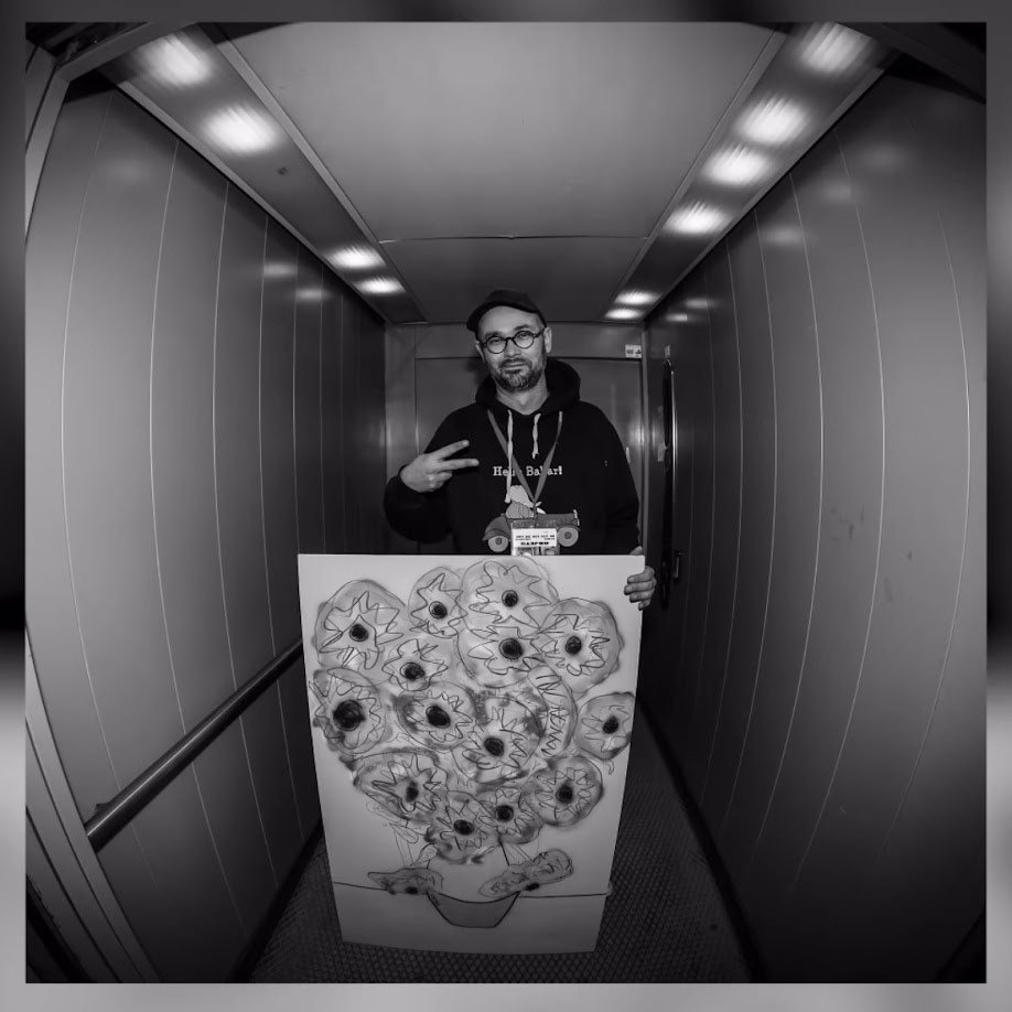 Portrait en noir et blanc de l'artiste dans un ascenseur, tenant un grand dessin de tournesols